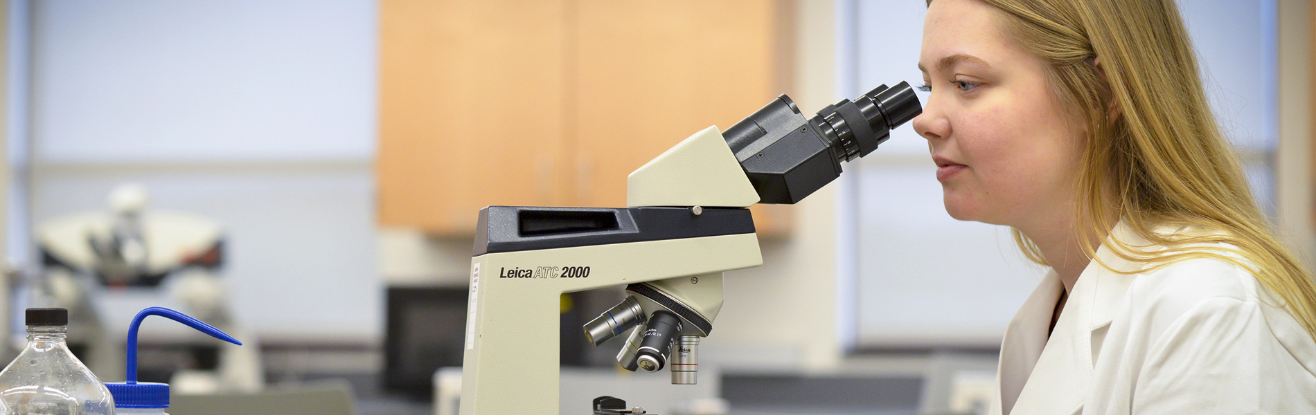 Female La Roche student wearing a white lab coat looking into a microscope.