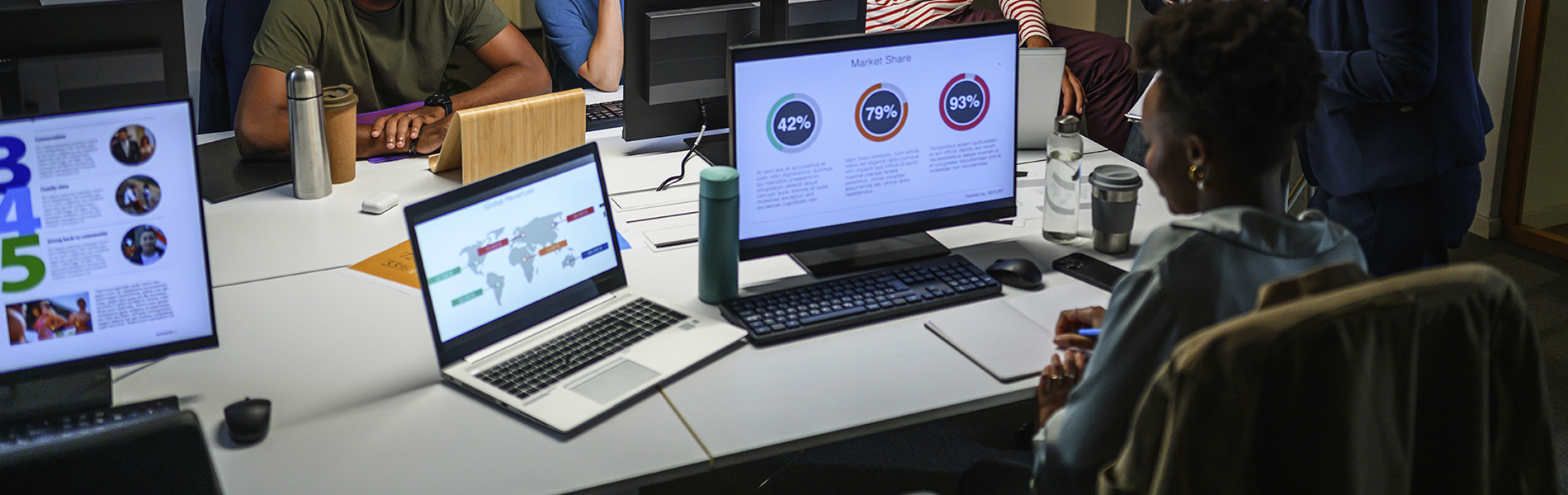 A person is seated at a desk, working on a laptop displaying a pie chart alongside another laptop showing a map and statistics. Another individual is seated nearby, also using a laptop.