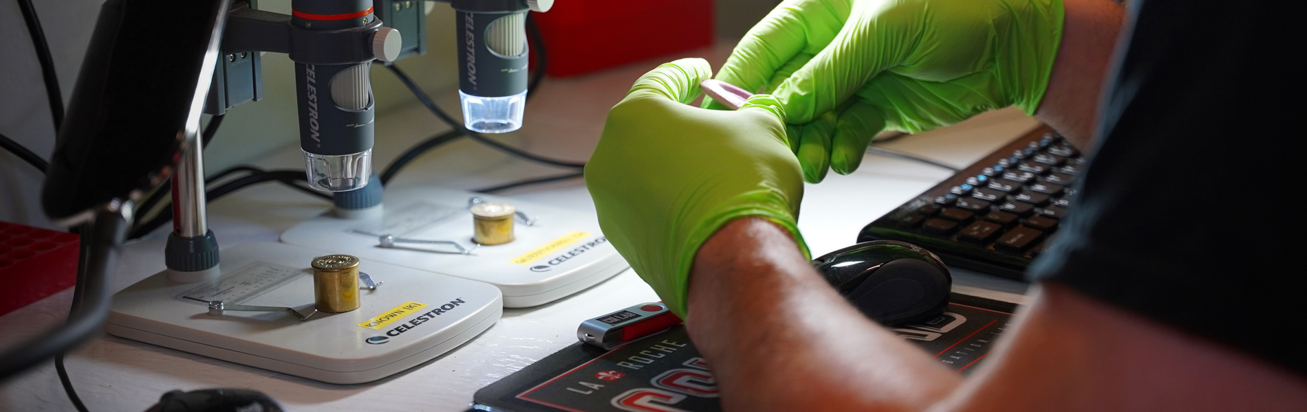 A person wearing green gloves working with a microscope in a laboratory setting with various tools and electronic equipment visible.