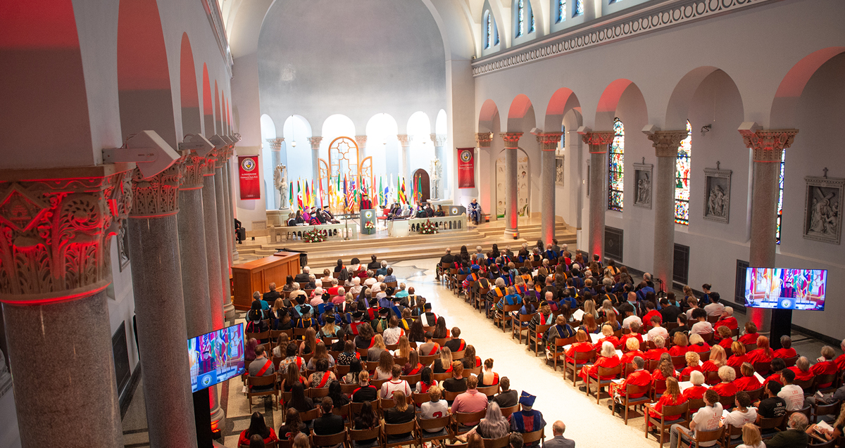 Interior of a crowded Mother of Divine Providence Chapel during Dr. Christina Clark's inauguration ceremony, with attendees seated and facing the altar where multiple clergy members are present. The church features arched columns and vibrant stained glass windows. Monitors display the ongoing ceremony for enhanced visibility.