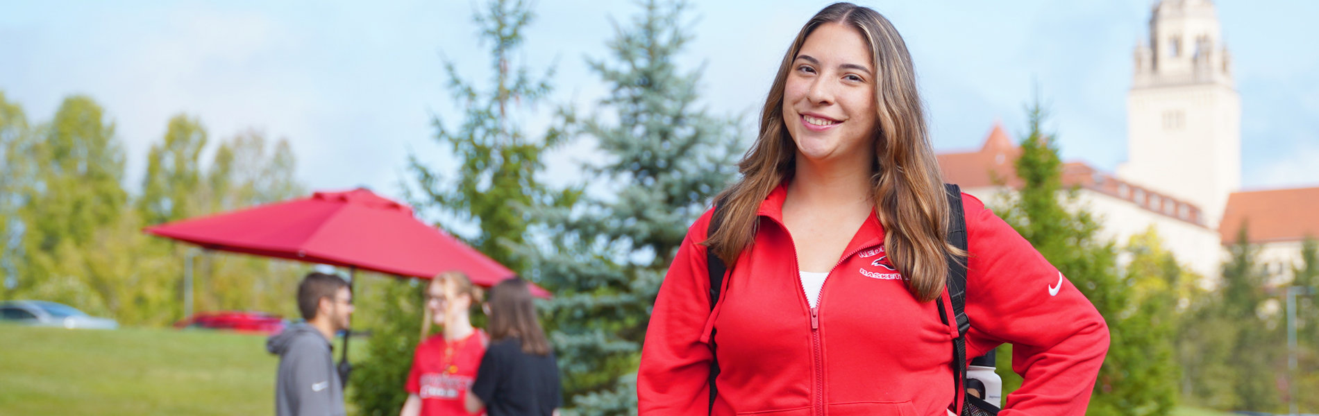 Female student at La Roche University. She’s standing in the foreground, smiling at the camera, wearing a red zip-up jacket and carrying a backpack