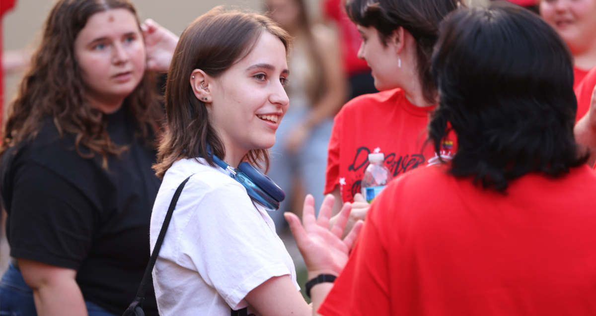 A person smiling and engaging in conversation with others at an outdoor gathering, with several people wearing red shirts in  background.