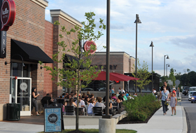 Groups of people dining outside at tables with red umbrellas at Hello Bistro. Two people walk on a sidewalk near the restaurant.