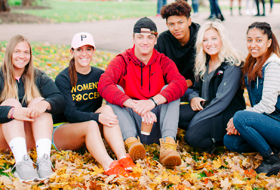 Six La Roche University students sit on campus on the lawn covered with fall leaves.