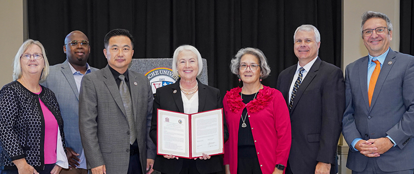 Group of seven individuals standing side by side in a conference room, holding a framed certificate. One person is displaying the certificate towards the camera. They are dressed in business attire.